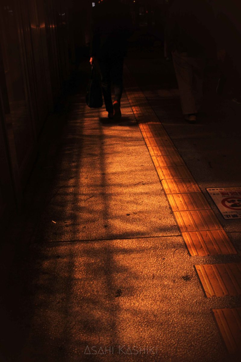 歩行者の足元の写真。店の明かりが交差して道に模様を作っている