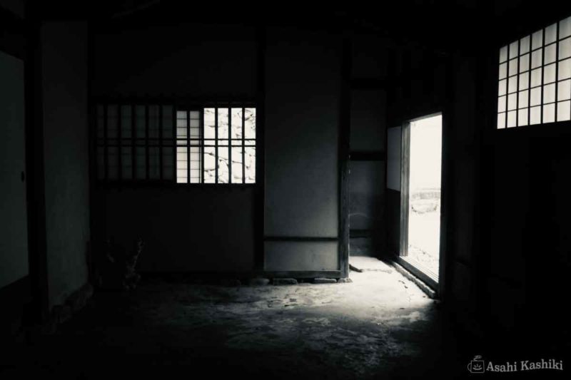 古いお屋敷の中の眺め。正面と右側に格子窓があり、右前には裏口への出口がある。室内は薄暗い。