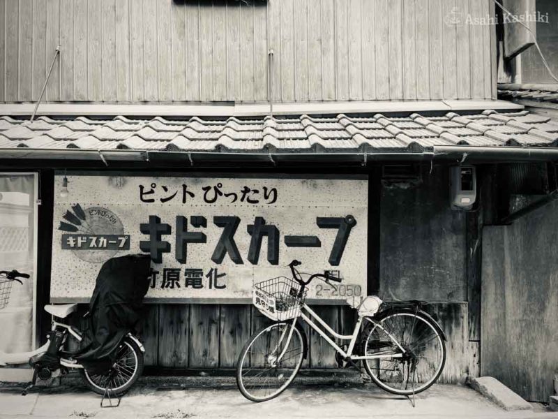 軒下に並ぶ自転車２台。壁には看板がかけられており、ピントぴったり、キドスカープ、竹原電化、と書かれている。
