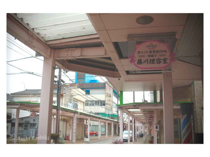 駅前の大きな商店街の一角。歩道に沿って屋根がかけられている。手前には「凛とした男前創作処 藤川理容室」の文字が見える。