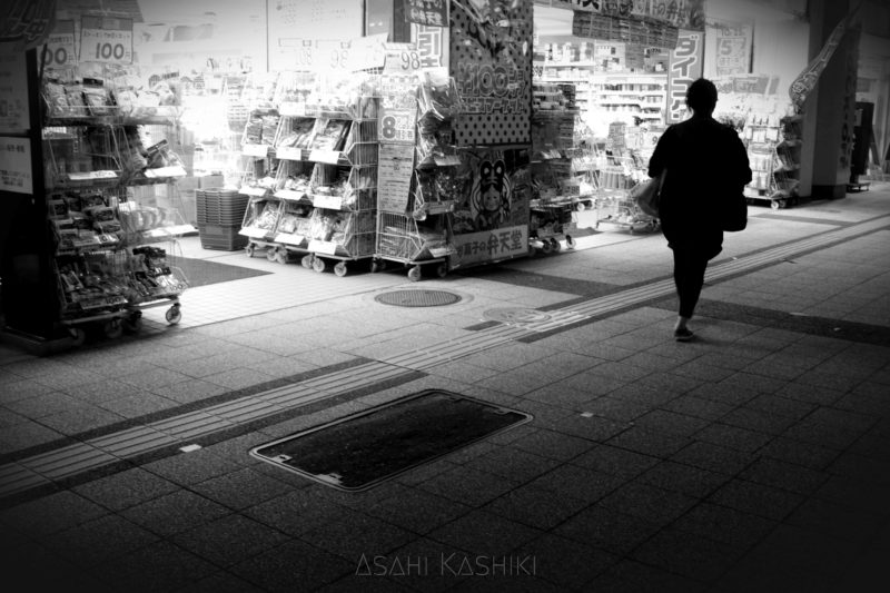 夜の薬局と、その前を歩く人のモノクロ写真。